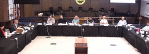 Nine people sit around a U-shaped conference table in a formal meeting room, with microphones and laptops in front and a seal on the wall behind them.