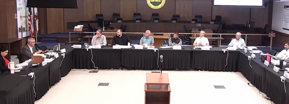 Nine people sit around a U-shaped conference table in a formal meeting room, with microphones and laptops in front and a seal on the wall behind them.